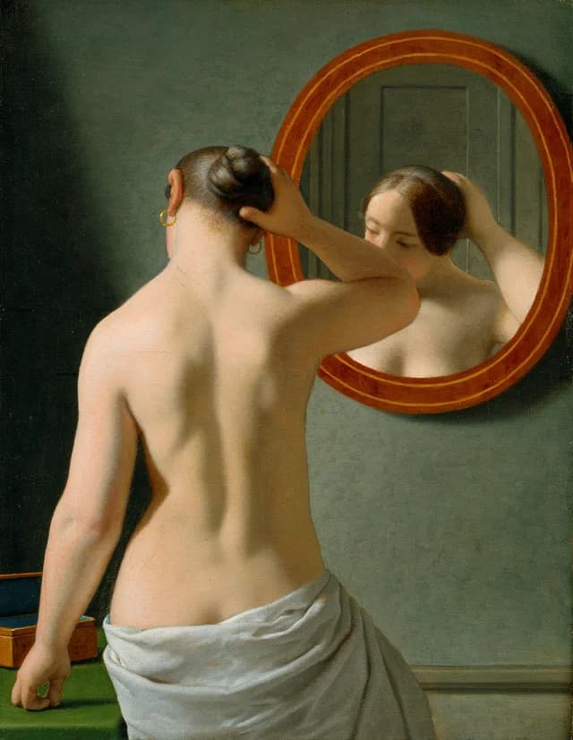 A nude woman doing her hair before a mirror